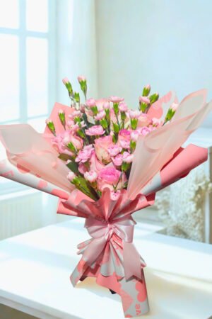 Ramo floral elegante en tonos rosados con rosas y mini claveles, envuelto en papel coreano translúcido y lazo satinado rosa.