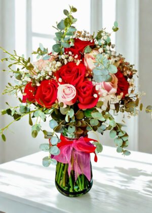 Arreglo floral elegante con rosas rojas y rosadas, flores delicadas blancas y follaje de eucalipto en florero de vidrio decorado con cinta rosada.
