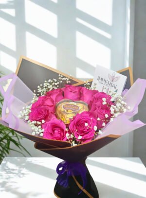 Bouquet elegante de rosas fucsias con bombones Valente en forma de corazón, gypsophila blanca y envoltura negra con detalles dorados.