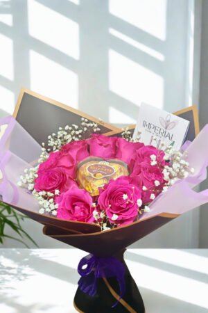 Bouquet elegante de rosas fucsias con bombones Valente en forma de corazón, gypsophila blanca y envoltura negra con detalles dorados.