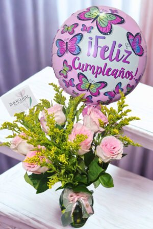 Arreglo floral de cumpleaños con rosas rosadas, solidago amarillo, florero de vidrio y globo decorativo de mariposas con mensaje Feliz Cumpleaños.